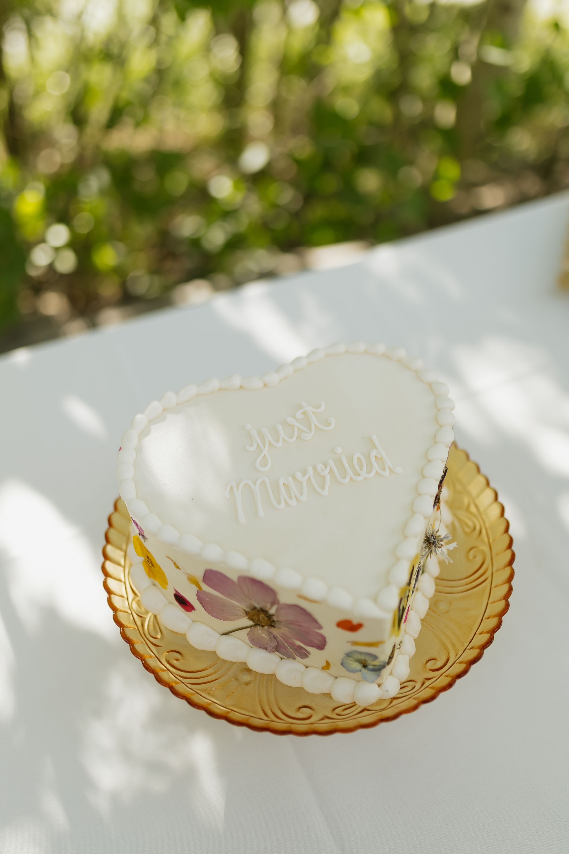 Heart-shaped white frosted cake with pressed flowers, on a gold plate, outdoors with 