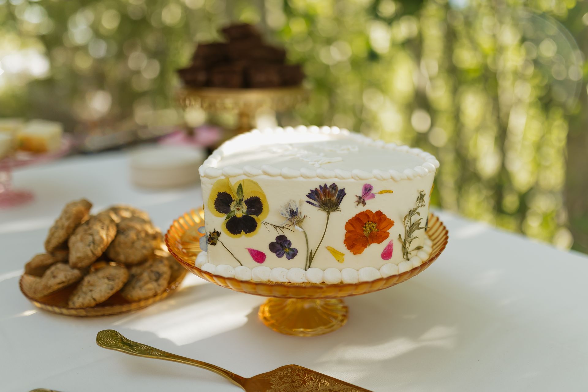 Cake decorated with pressed flowers on a gold cake stand, cookies and other treats on a white table.