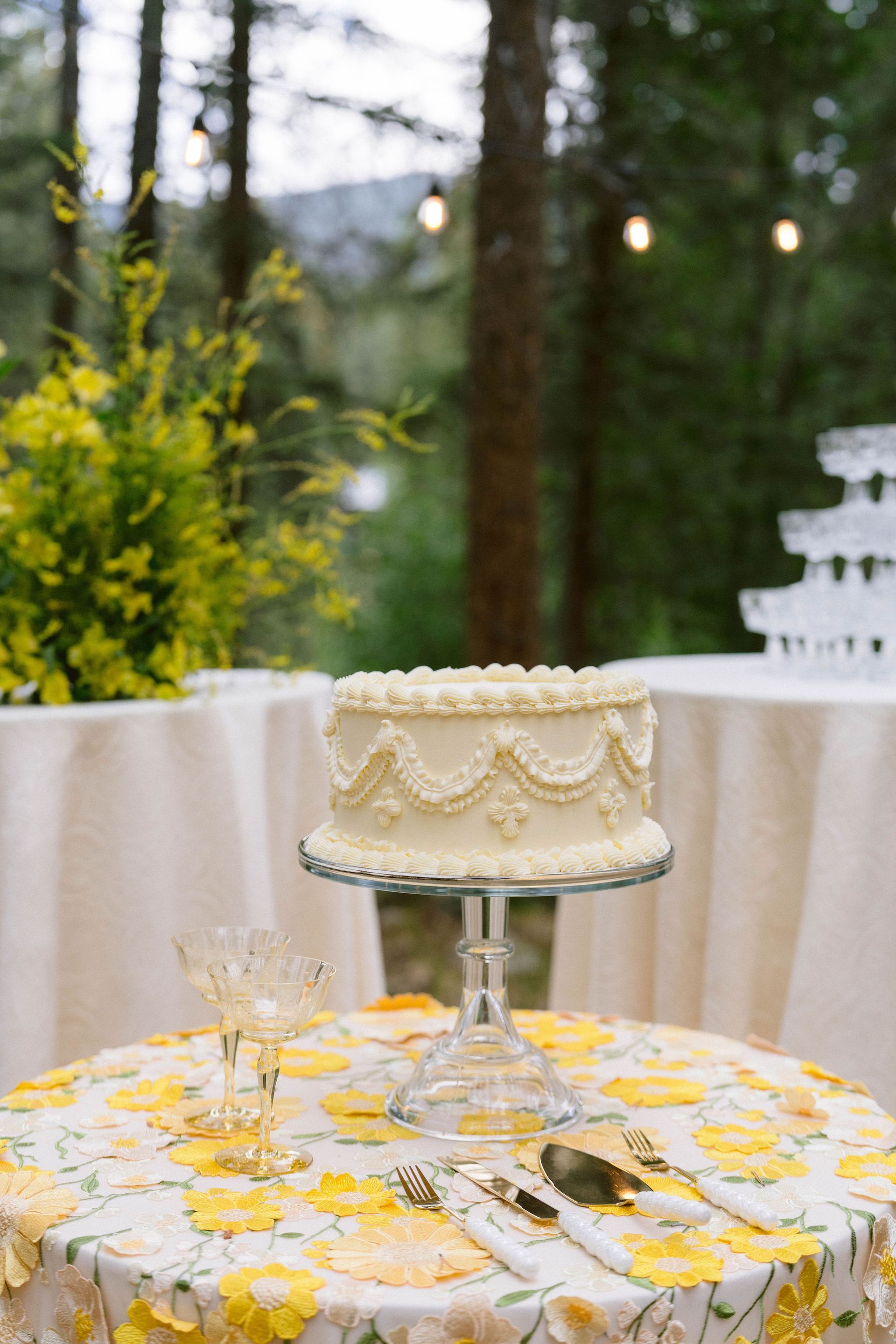 Wedding cake on glass stand, outdoor setting, yellow floral tablecloth.