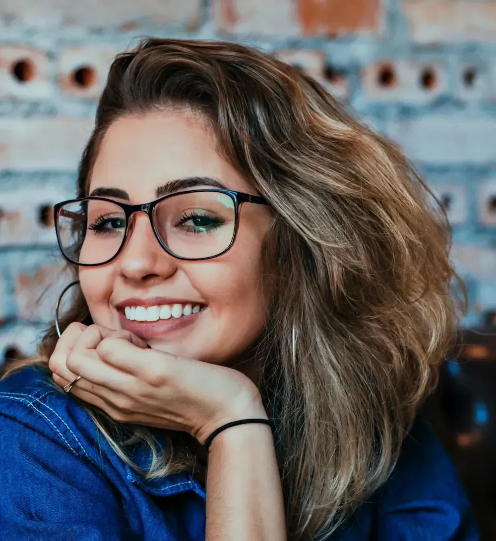 En smilende person med skulderlangt hår og briller hviler haken på hånden mot en murvegg i bakgrunnen.