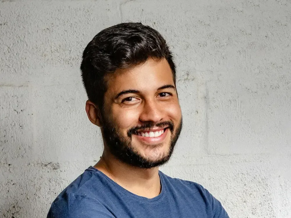 En person med kort, mørkt, krøllete hår og skjegg som smiler varmt til kameraet, iført en blå t-skjorte mot en grå vegg.