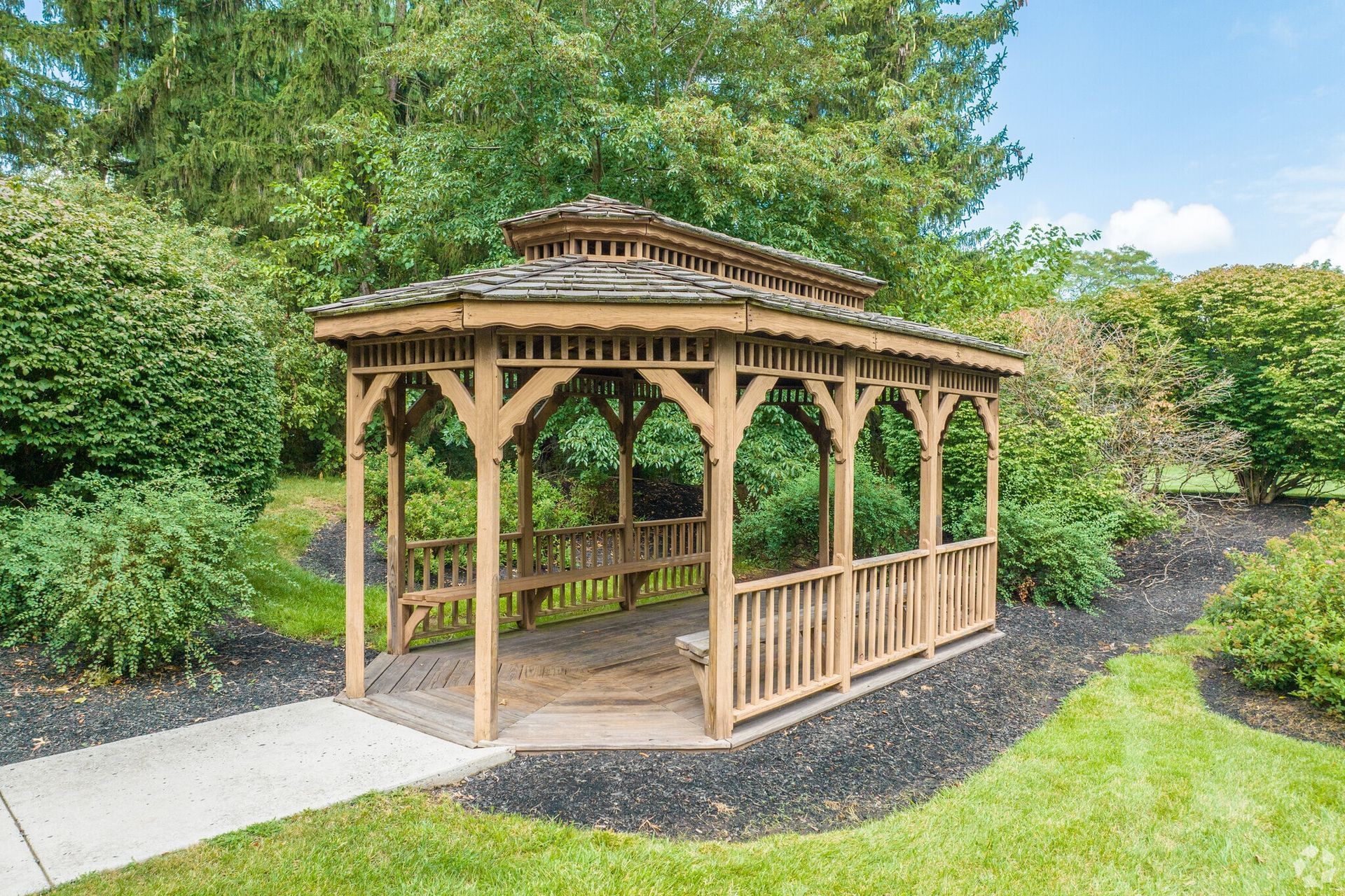 Wooden gazebo with benches, in a garden setting, surrounded by greenery and a paved path.