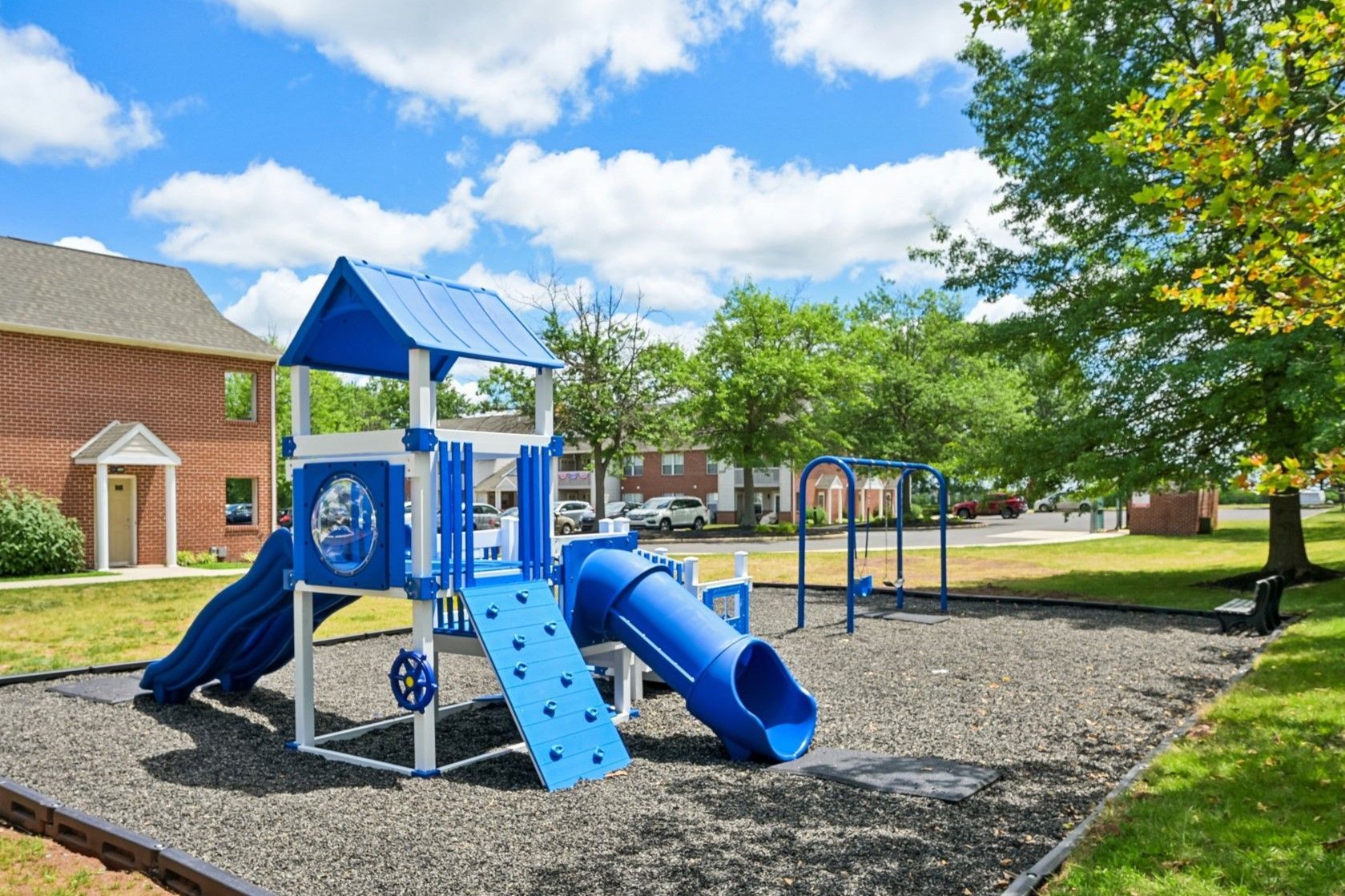 Blue playground climber and slide on a mulch play area beside brick buildings under a sunny sky.