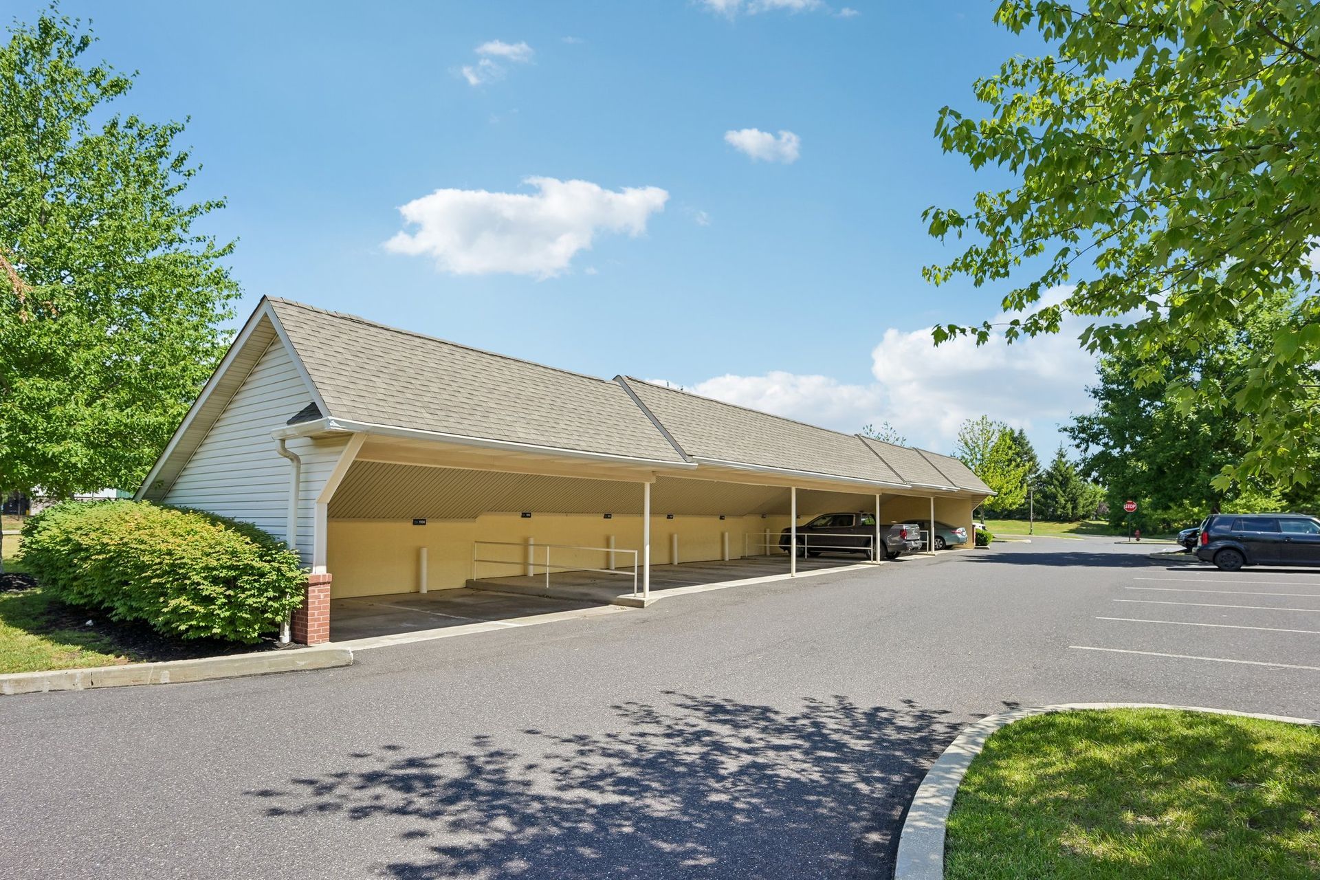 Covered parking area beside a brick building with trees and a paved road under a blue sky