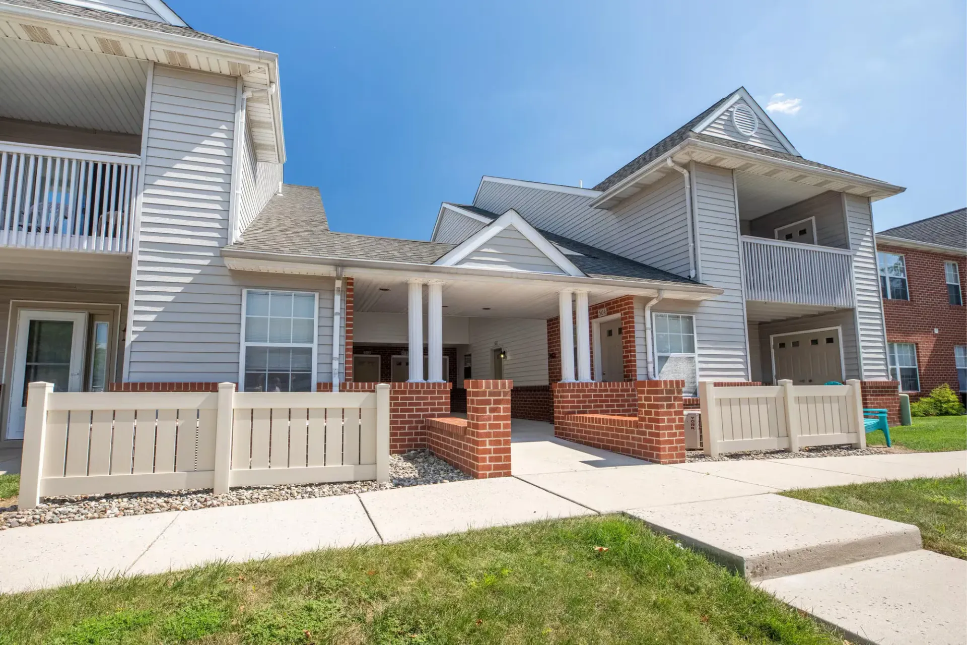 Wooden gazebo with benches, in a garden setting, surrounded by greenery and a paved path.