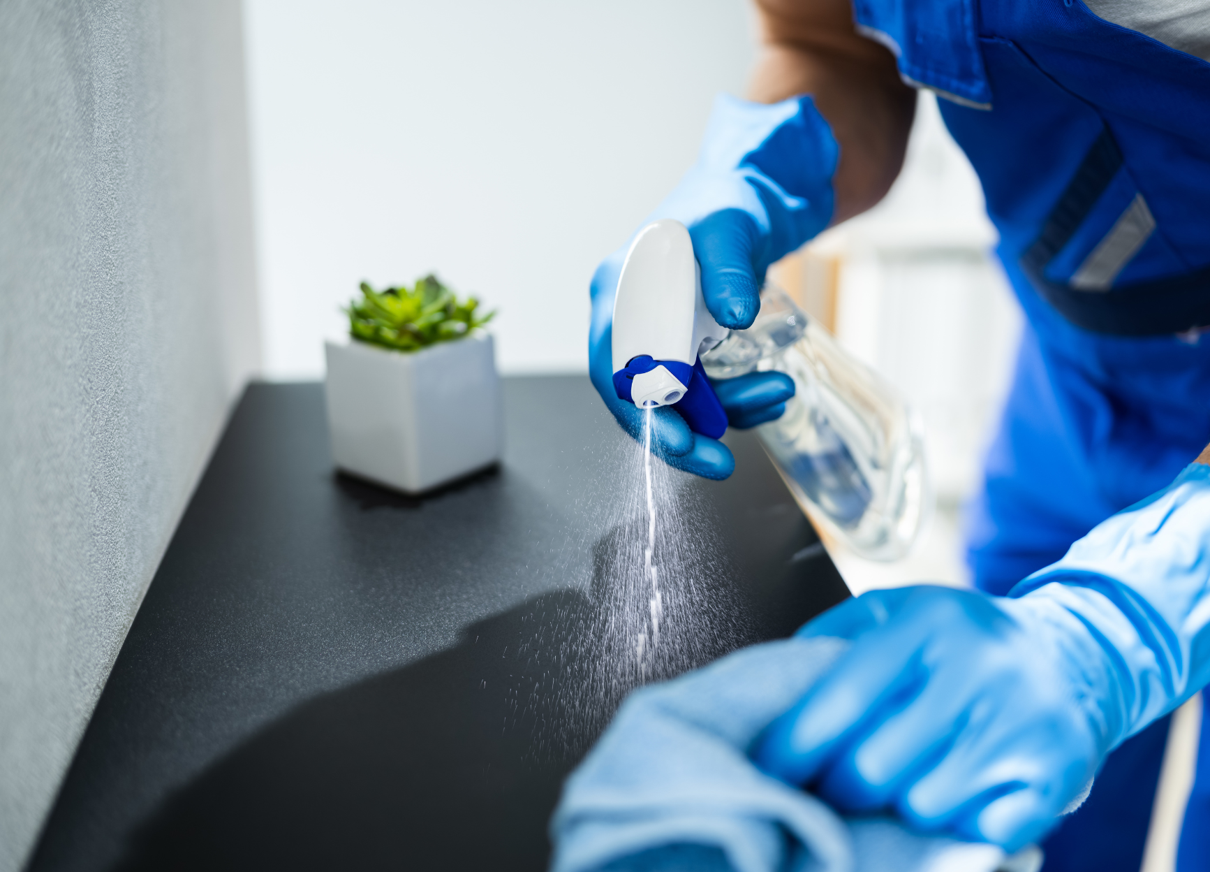A person wearing blue gloves is cleaning a sink with a cloth and spray bottle.