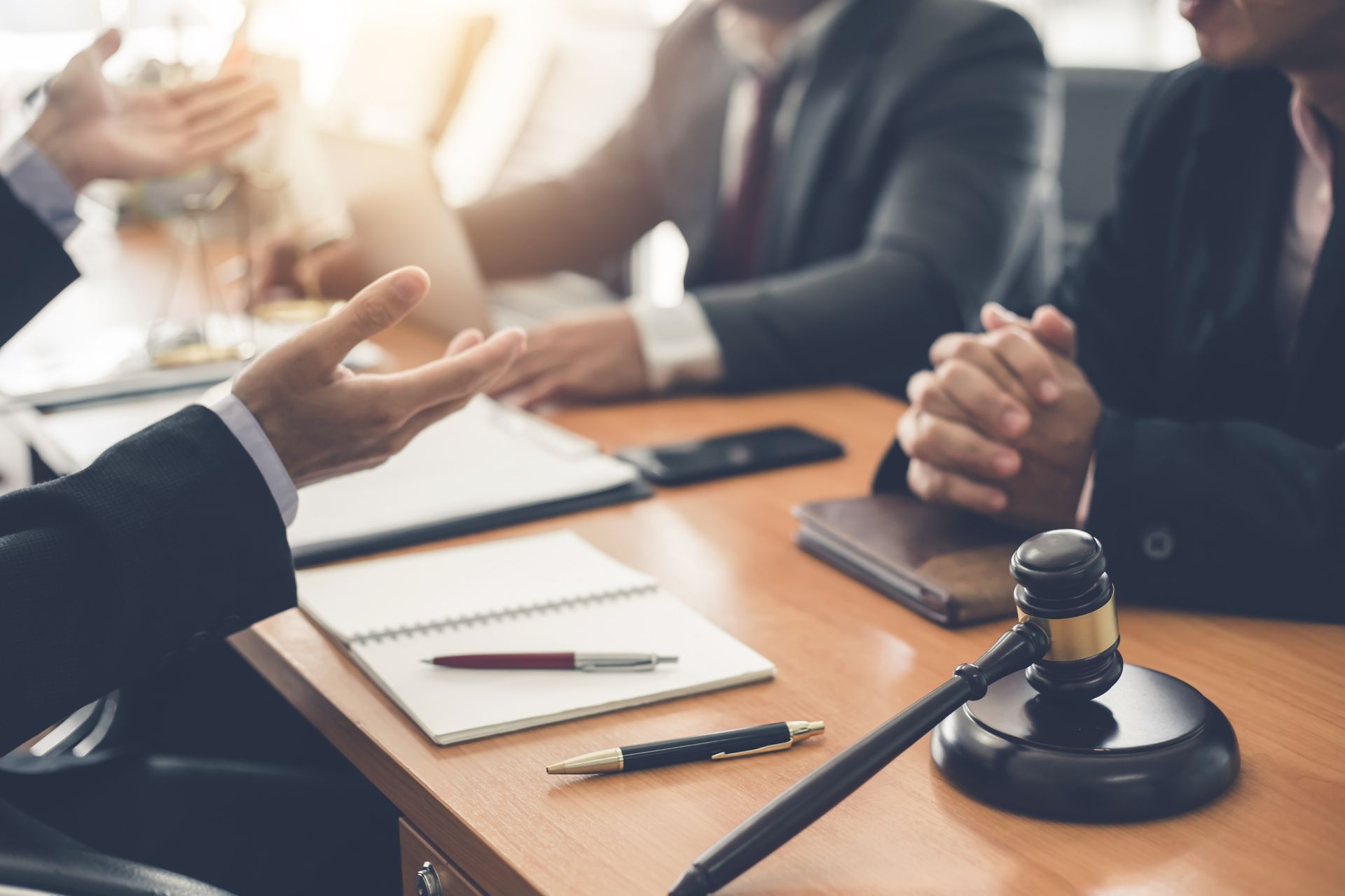 Legal professionals in suits sit at a desk with notebooks, a pen, and a judge's gavel during a meeting.