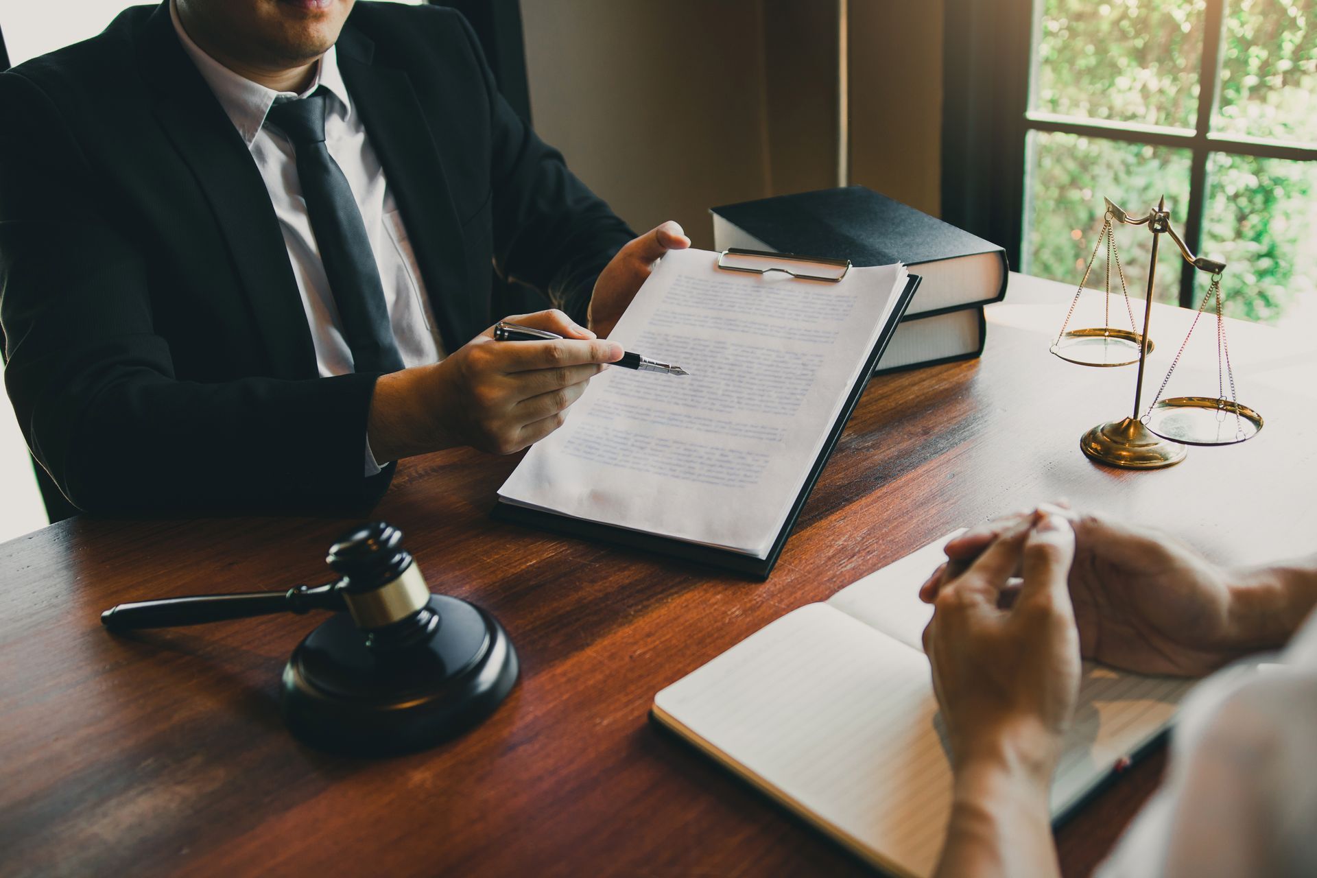 Two professionals sit at a desk with an open book, a gavel, and a balance scale in a legal or office setting.