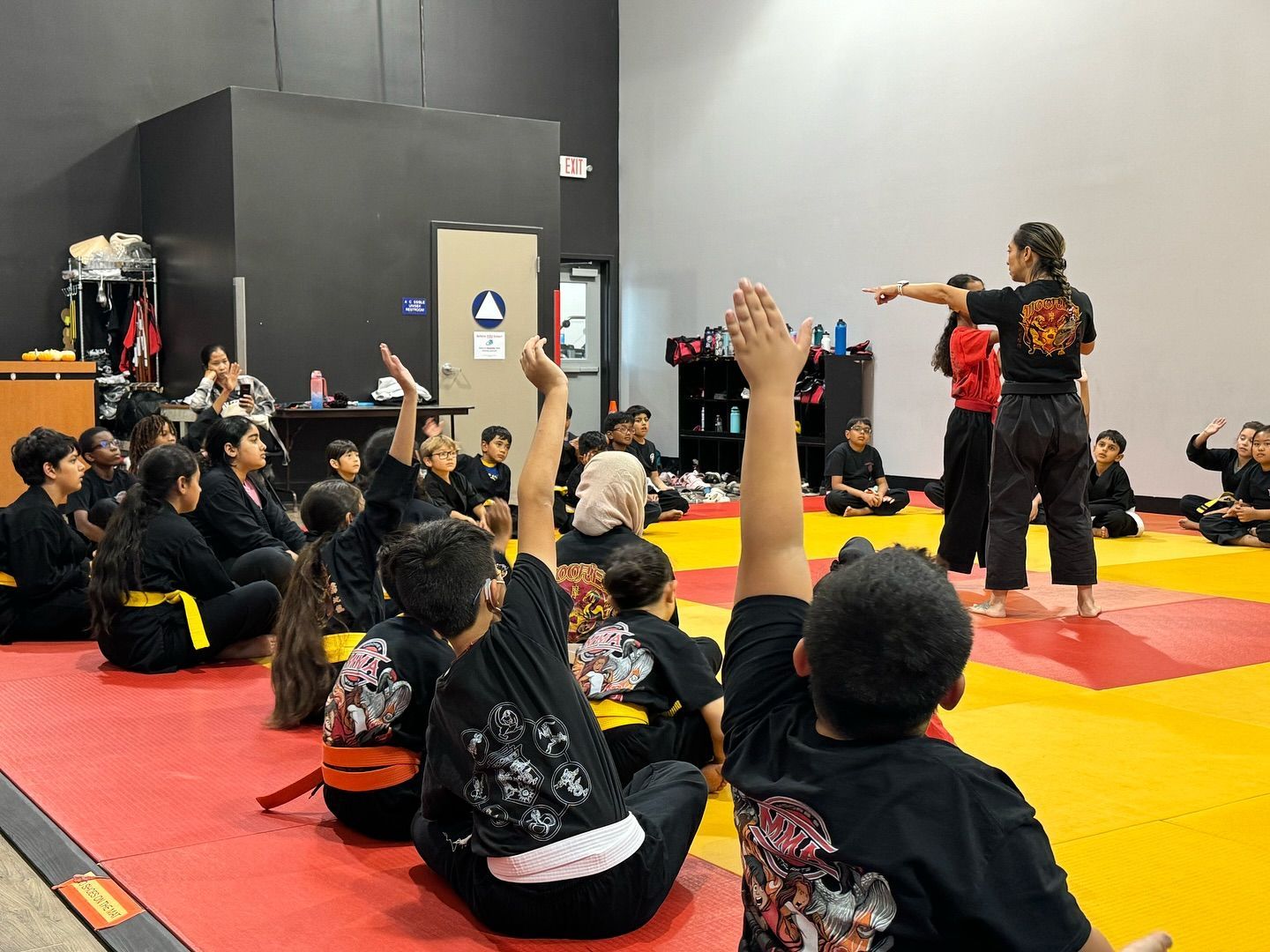 A group of children are sitting on the floor raising their hands to answer a question.
