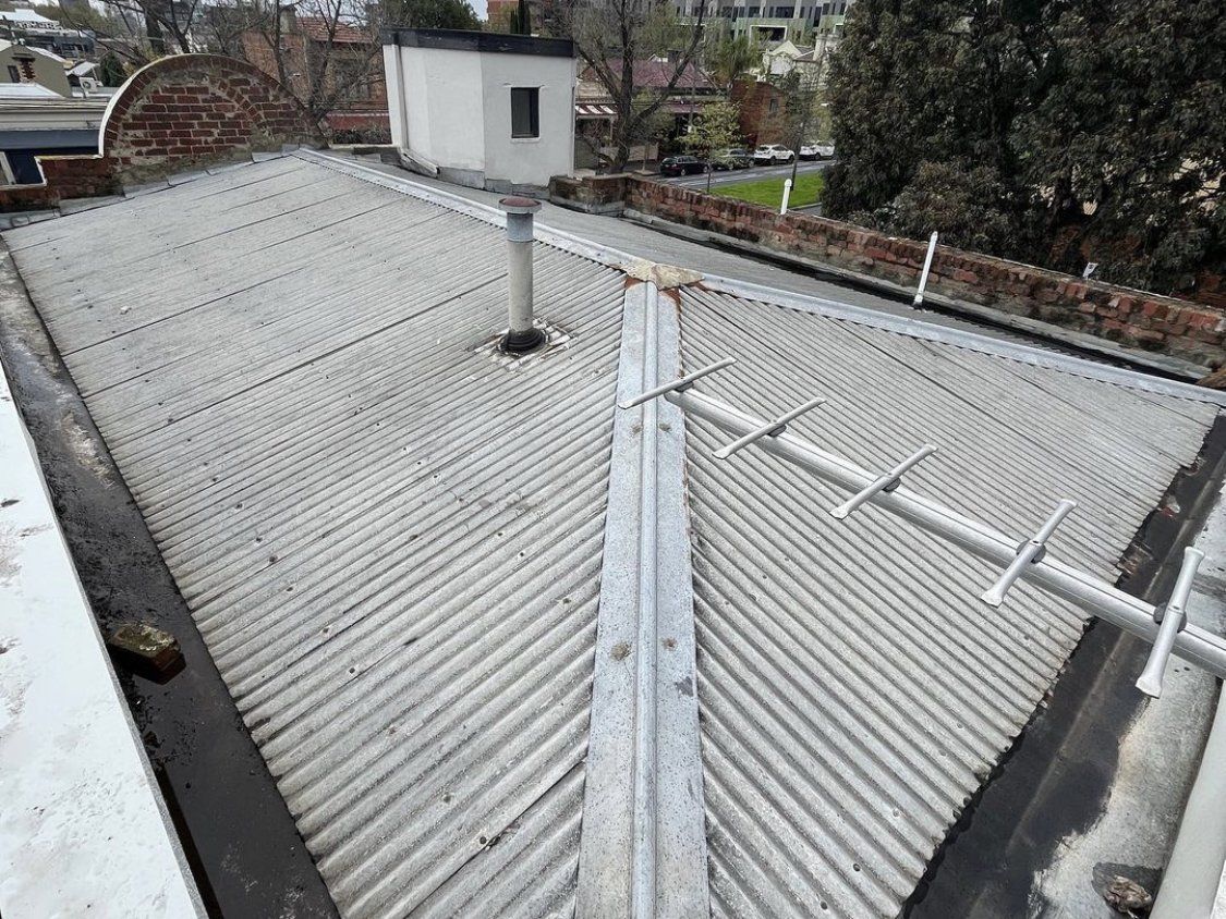Image depicts a very old corrugated iron roof that is rusted and full of leaks. The roofline is surrounded by a brick parapet wall which holds box guttering in place around the entire roof. The box guttering is also leaking.