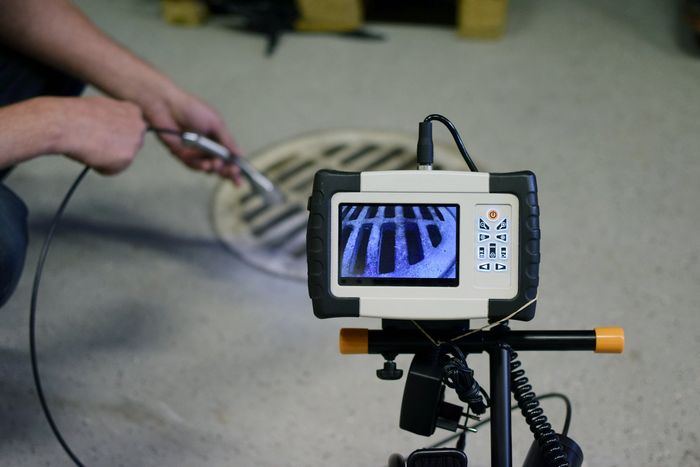 Image depicts a concrete draw with a drain grate. A man is holding a wired camera and directing it down the drain. The camera body is fixed to a tripod and the imagery is streamed back to the hard drive so the worker can down the drain and identify any obstructions.