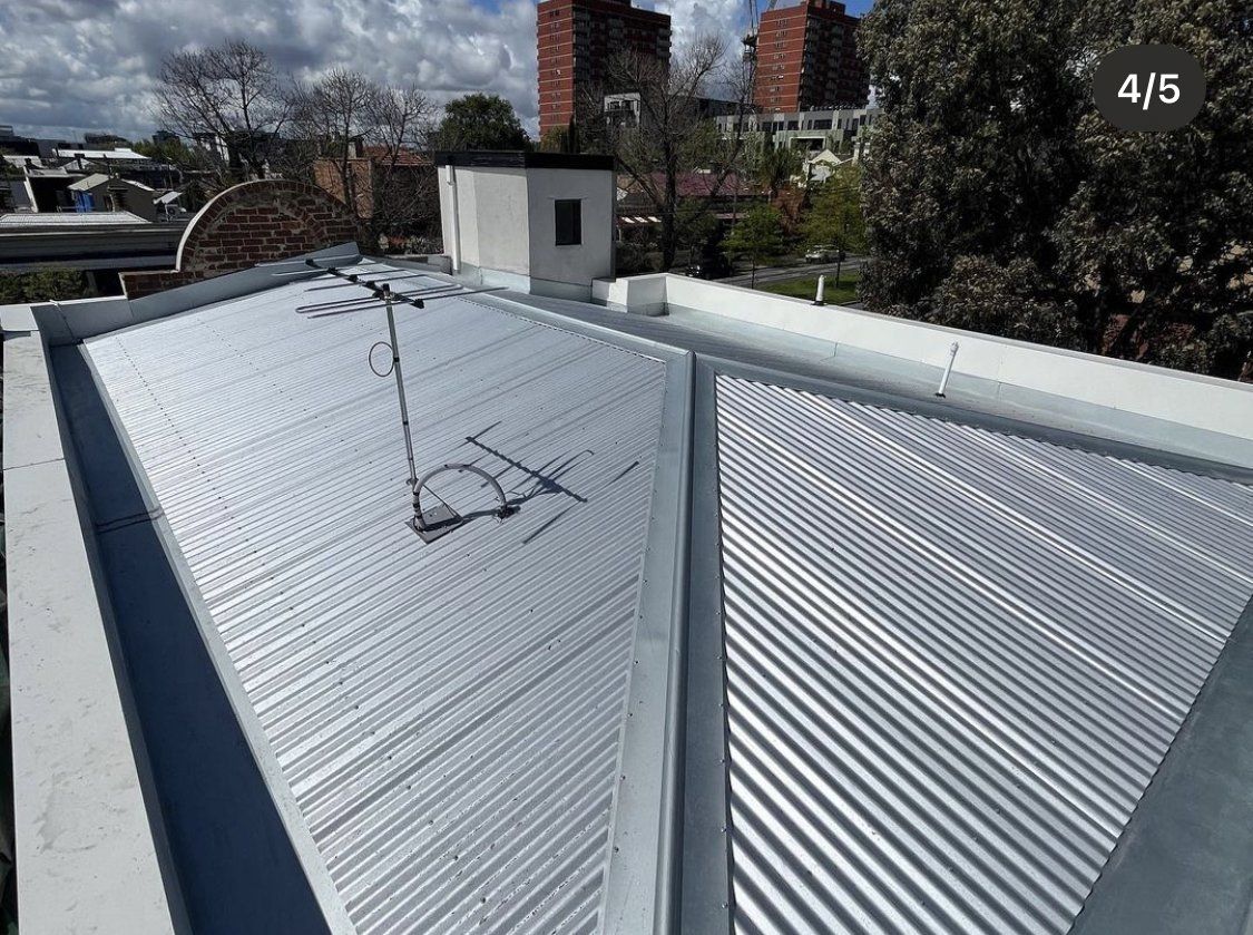 Image depicts a brand new corrugated iron roof that replaced an old leaking roof. The roofing structure is surrounded by brick parapet wall which has box guttering around the entire roofline.