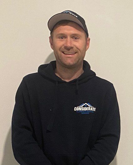 The image depicts Considerate Plumbing & Roofing director Tim Birmingham in a black hoodie with the logo Considerate Plumbing & Roofing on the left side. He is standing against a wall for a portrait shot with a black Considerate Plumbing & Roofing peak cap on.