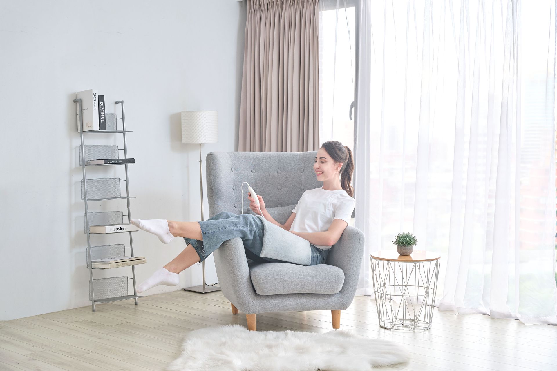 Woman relaxing in gray armchair, smiling while using a tablet in bright living room.