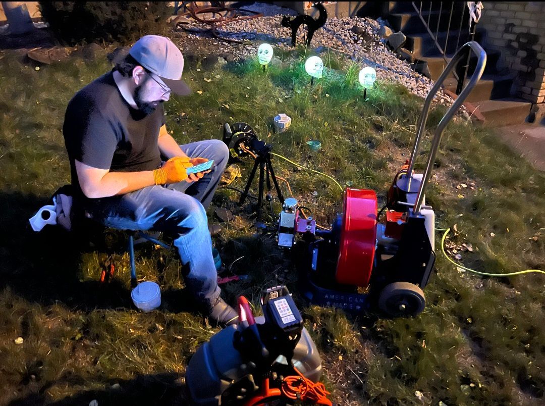 Man with remote controls lawn mower in a yard decorated with skull lights at night.