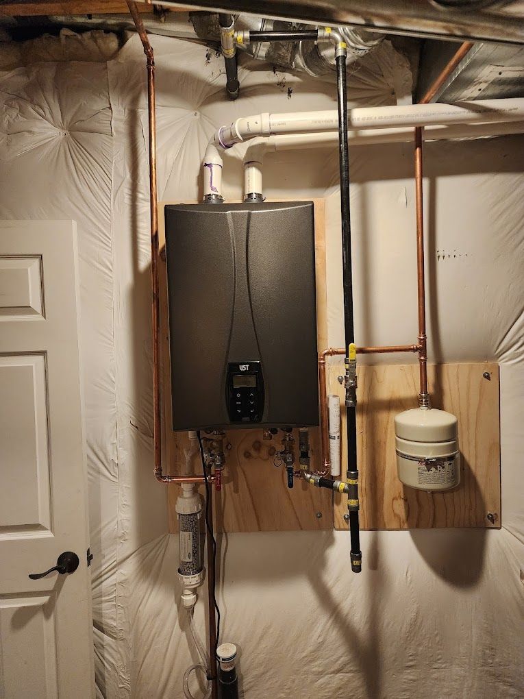 Wall-mounted, dark metallic water heater with copper piping, installed in a basement near a door.