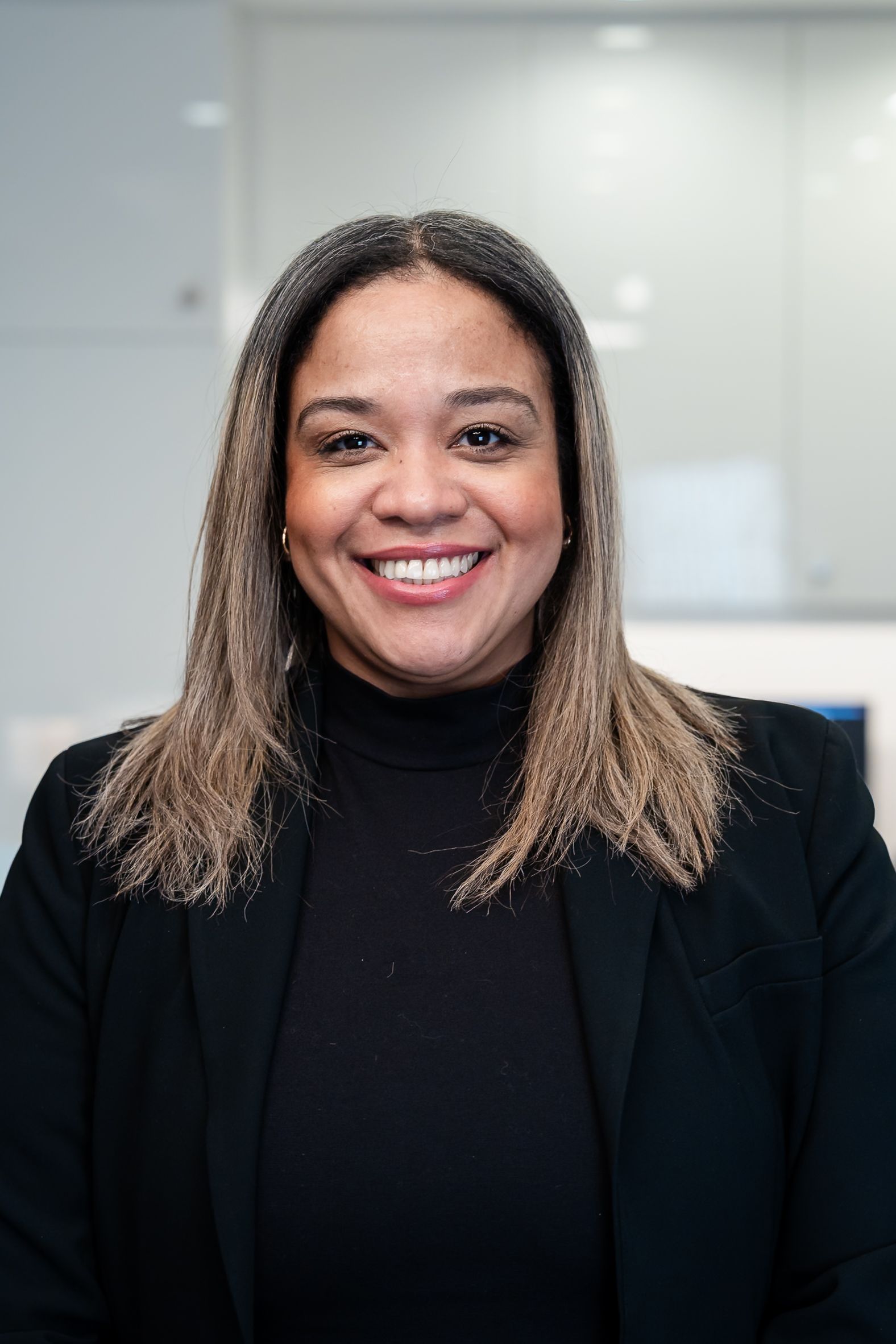 Woman in blazer smiling, standing in white hallway with arms crossed.