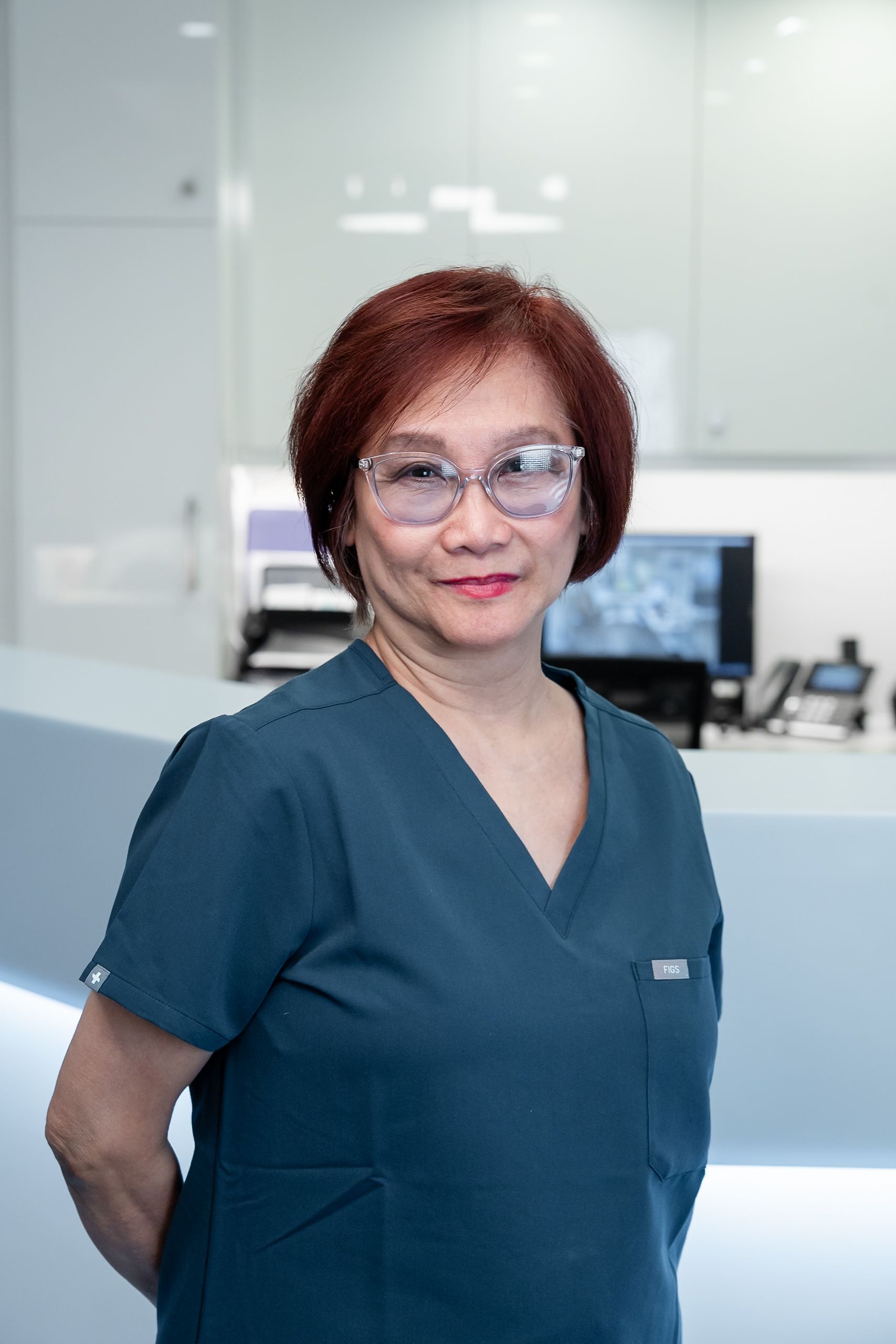 Woman in black scrubs, hands on hips, smiling in a white hallway with a bright overhead light.