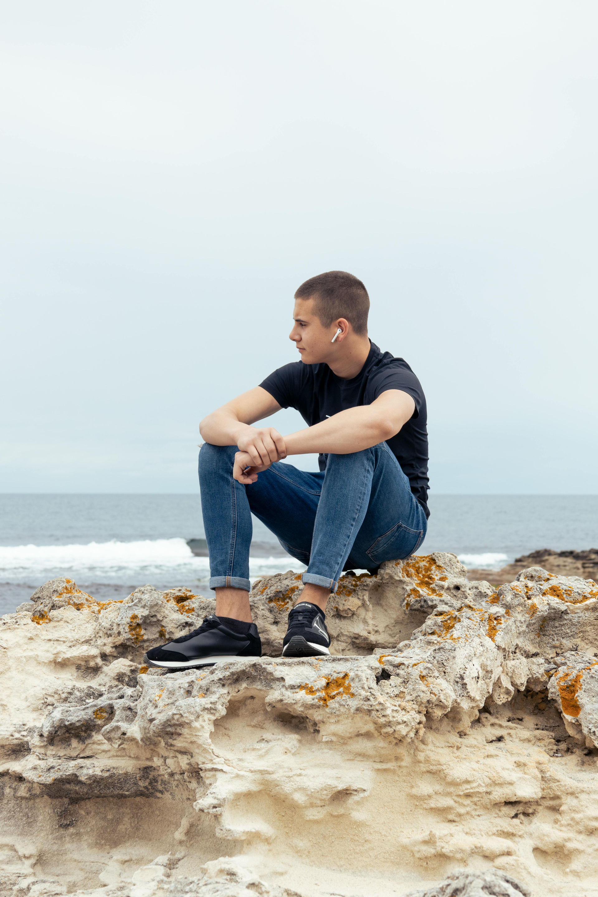 Man zit op rotsen bij de zee en kijkt weg. Hij draagt ​​een spijkerbroek, een zwart shirt en sneakers.