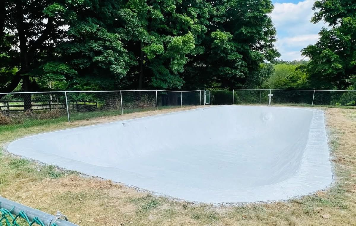 A large white swimming pool is surrounded by trees and a fence.