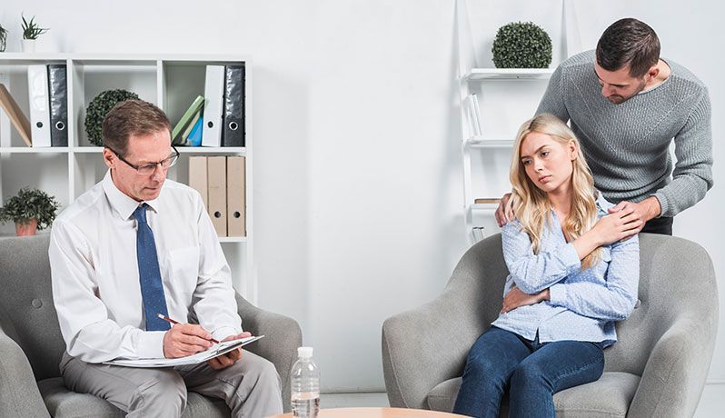 A woman is writing on a clipboard while a man and woman sit on a couch.
