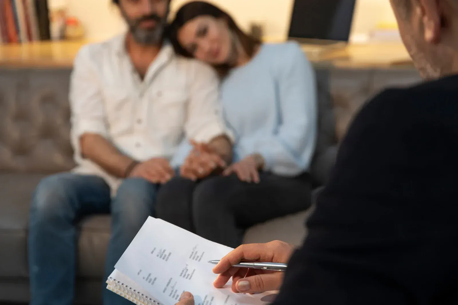 A man and a woman are sitting on a couch while a person writes on a piece of paper.