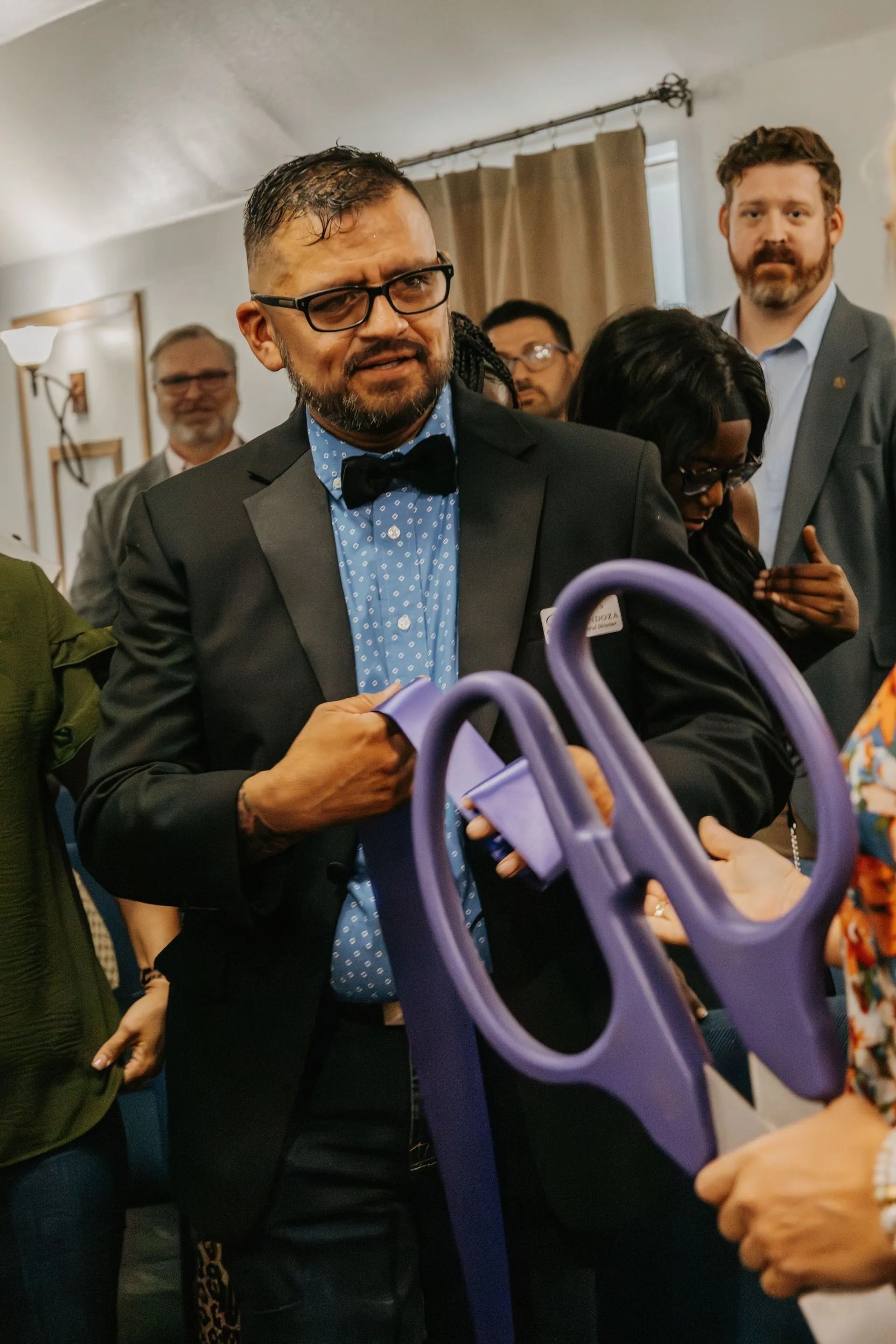 A man in a suit and bow tie is holding a pair of purple scissors.
