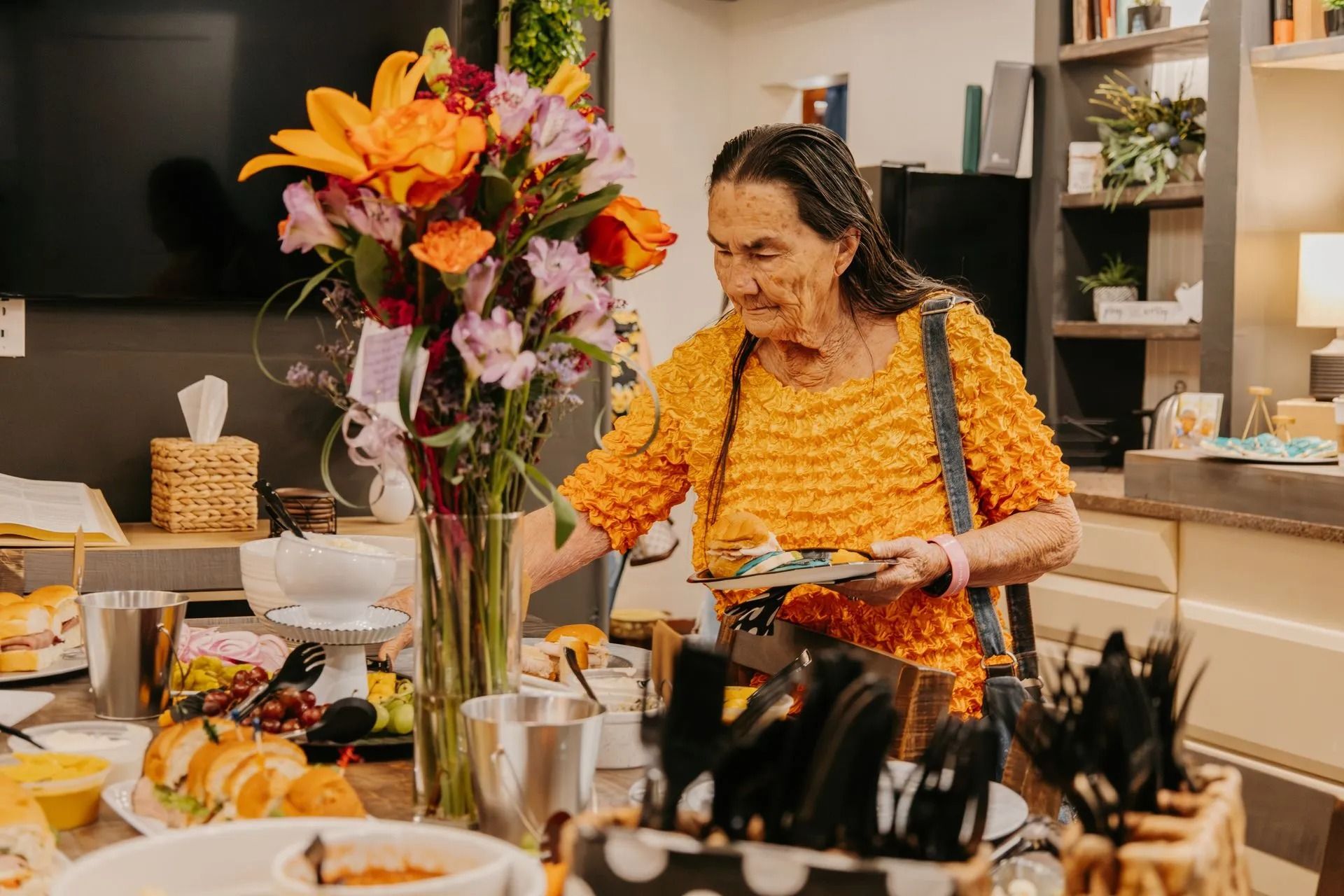 An elderly woman is standing at a table with a vase of flowers.