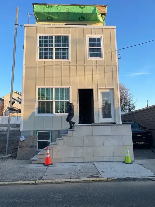 Two-story house under construction with worker on front steps and orange cones on snowy driveway.