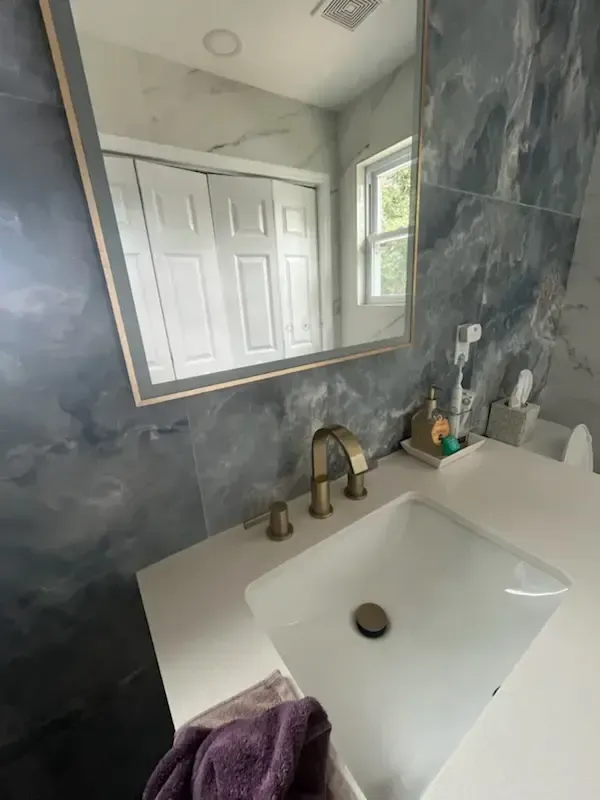 Bathroom vanity with white sink, brass faucet, gray marble wall, and mirror reflecting a door and window