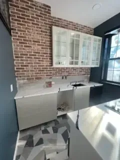 Modern kitchen corner with brick accent wall, white cabinets, gray lower cabinets, and geometric tile floor.