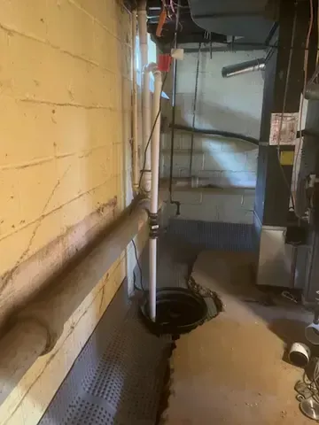 Basement utility area with yellow wall, pipes, and a sump pump opening on the floor.