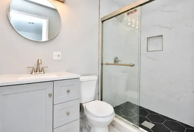 Modern white bathroom with vanity, round mirror, toilet, and glass shower enclosure with black tile floor accents