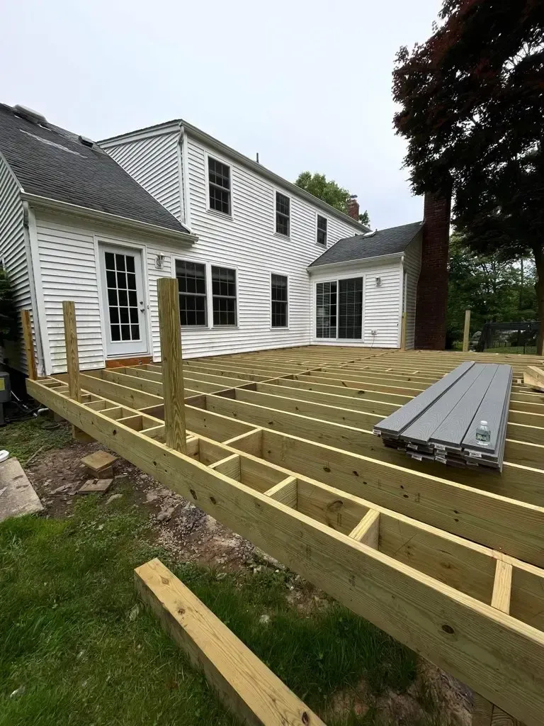 Deck framing under construction beside a white house, with lumber and joists laid out in the yard.