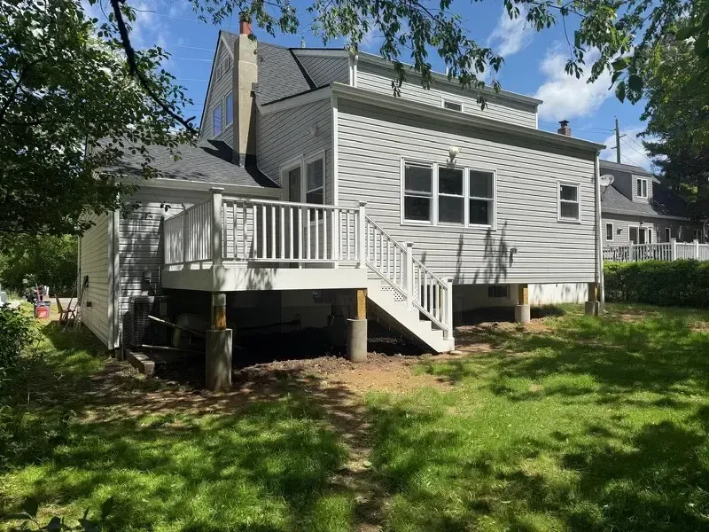 White house with a raised deck and stairs in a grassy yard under a sunny sky.