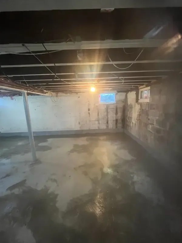 Empty unfinished basement with exposed ceiling beams, concrete floor, and a bright light near a small blue window.