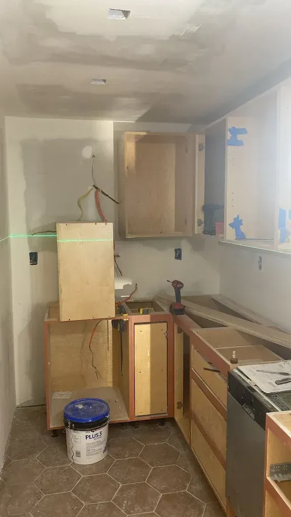 Narrow kitchen under renovation with cabinets, tools, and paint buckets on the floor.