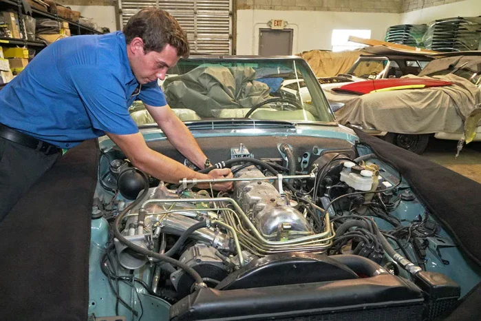 Mechanic in blue shirt working on the engine of a teal vintage car inside a garage.