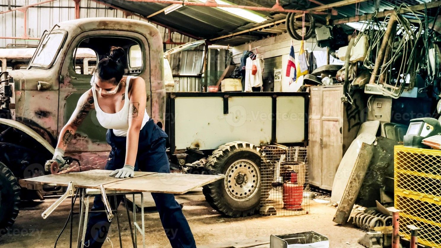 Woman in work clothes cutting wood, working on a vintage truck in a cluttered garage.