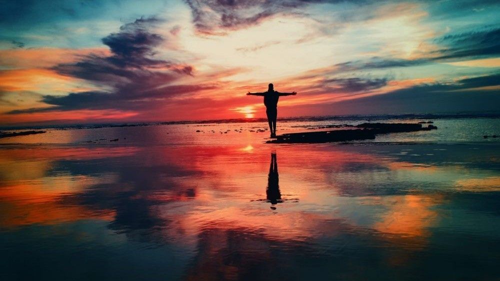 Silhouette of person with arms outstretched, facing vibrant sunset over reflective water.