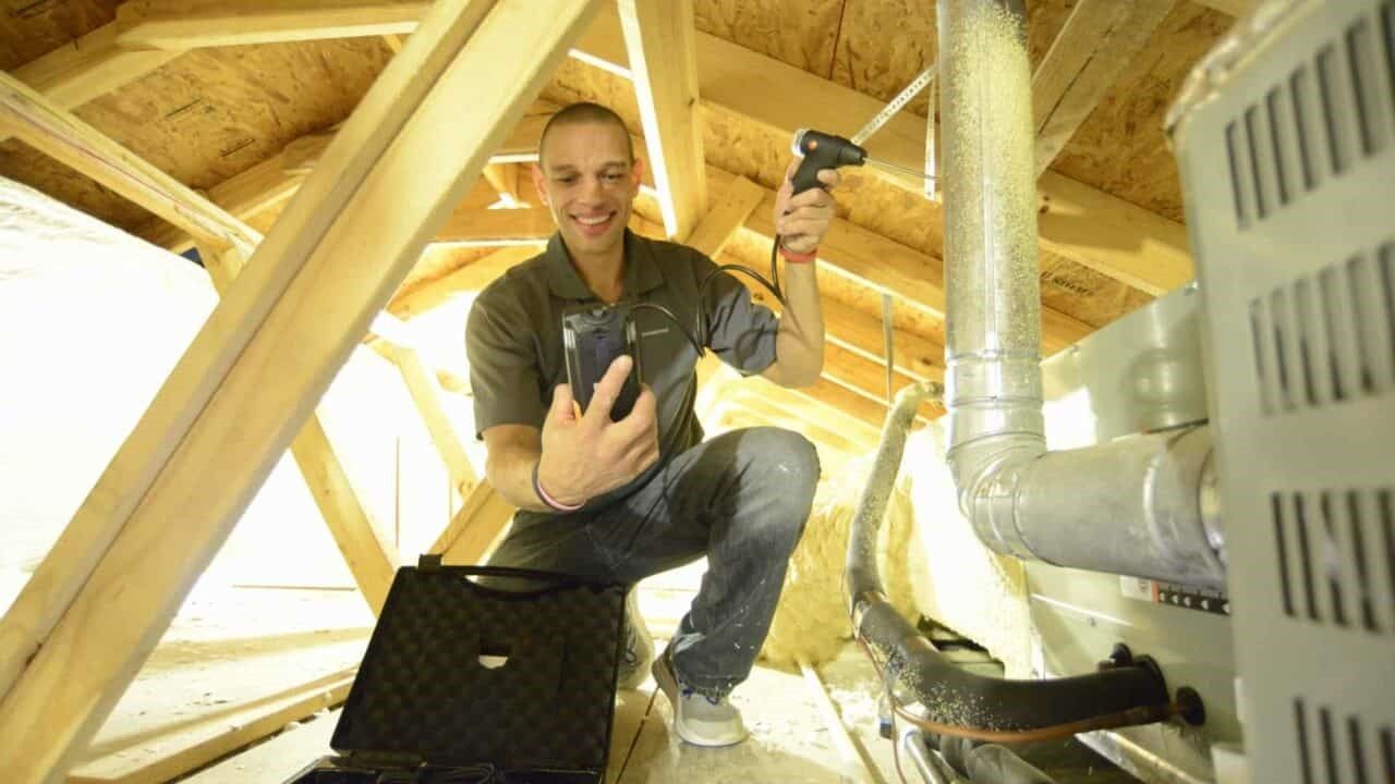 A man is kneeling down in an attic holding a tablet.