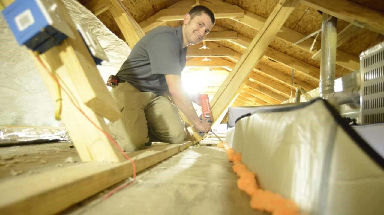 A man is kneeling down in an attic white applying foam sealant