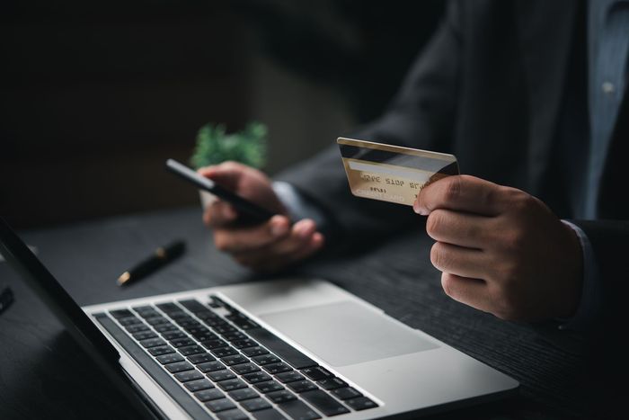 Hands holding a credit card and smartphone beside a laptop, suggesting online shopping or payment.