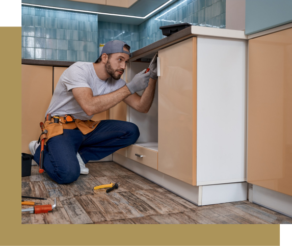 Man in work clothes fixing kitchen cabinet, kneeling with tools.