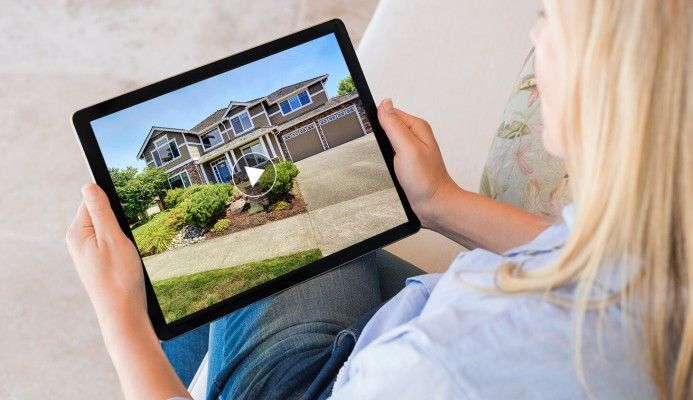 A woman is sitting on a couch holding a tablet with a picture of a house on it.