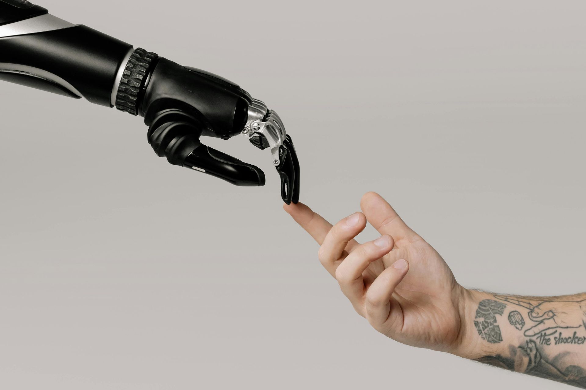 Black robotic hand touches a human finger. Gray background, tattoo on forearm.