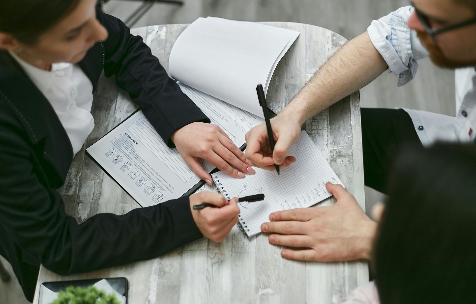 People at a table writing with pens on paper; business meeting setting.