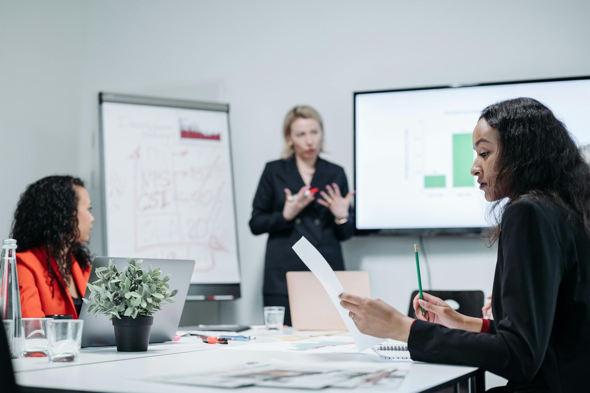 Businesswomen in meeting; one presents data, others listen, and work on laptops.
