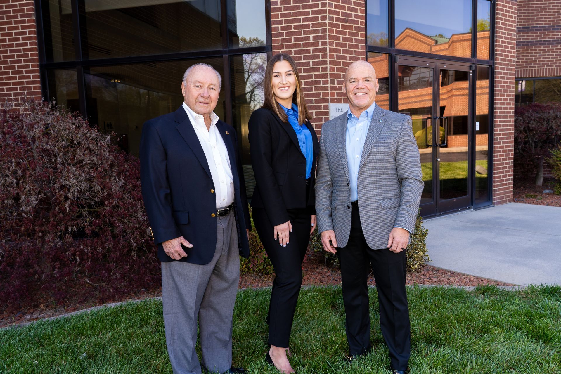 Three people standing outside a brick building. One wears a navy blazer, another a black suit, and the third a gray blazer.