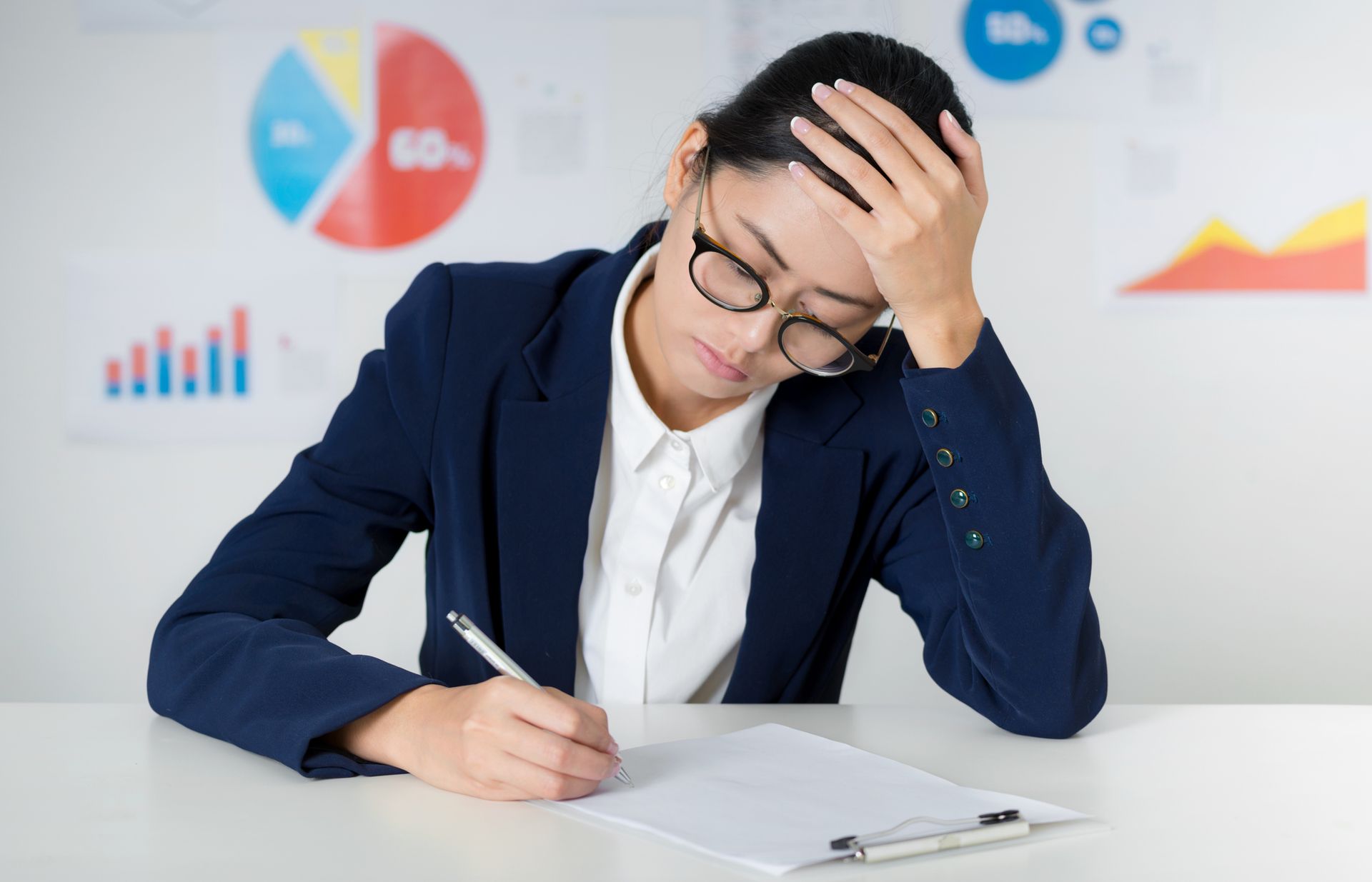 Woman in a suit, eyeglasses, and hand on forehead, writing on paper with data charts in the background, appearing stressed.